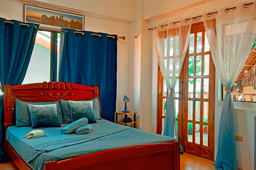 Opposite angle view of the comfortable queen bed, vibrant blue decor, and woodgrain balcony doors in the VIP Executive Suite San Carlos resort.